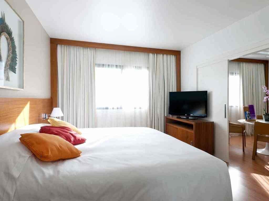 A hotel room with a bed, a television, a desk with a chair, and a window with curtains.