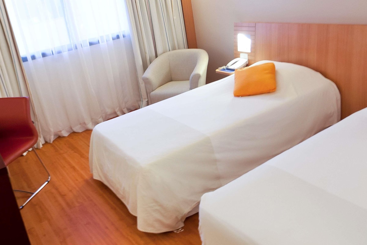 A hotel room with two beds, a chair, a desk with a phone, and a window with curtains.