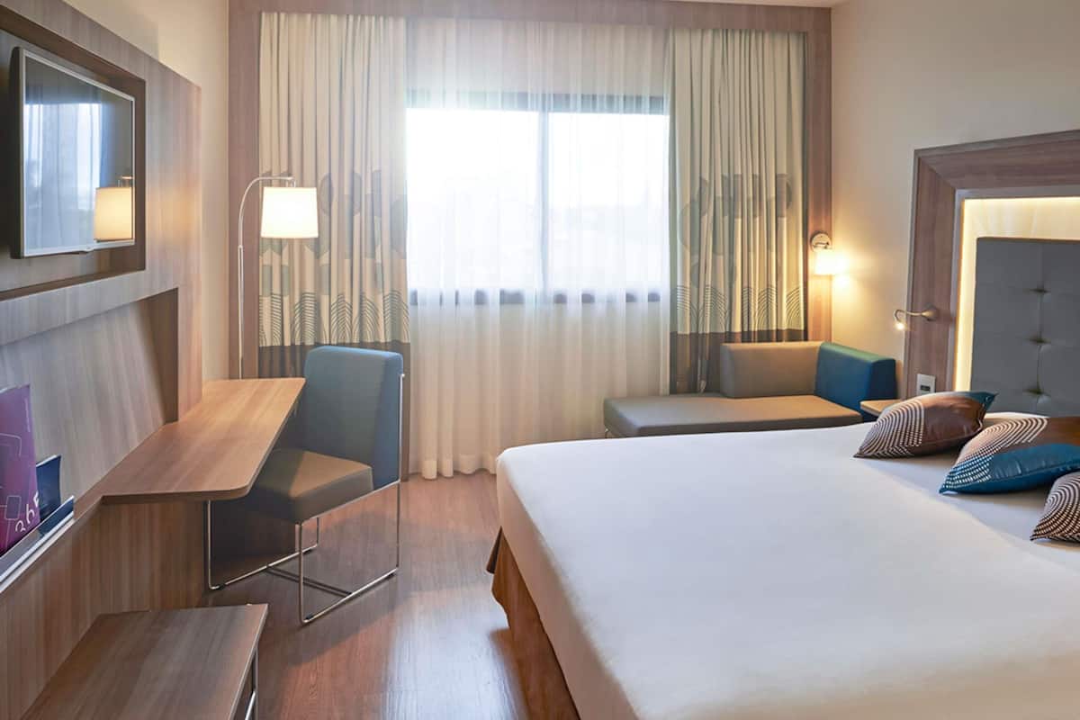 A hotel room with a bed, a desk, a chair, a TV, and a window with curtains.