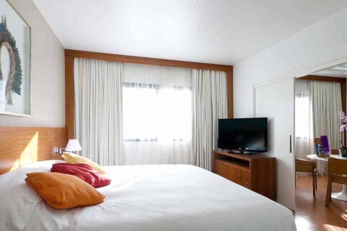 Quarto de hotel com cama, televisão, escrivaninha com cadeira e janela com cortinas.