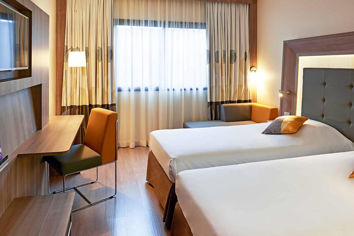 A hotel room with two beds, a desk, a chair, a TV, and a large window with curtains.