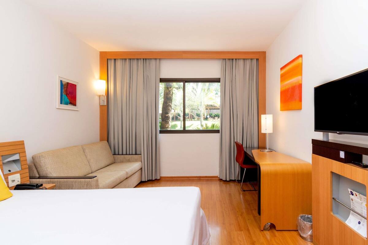 A hotel room with a bed, a desk, a TV, a chair, a lamp, and a window with curtains.