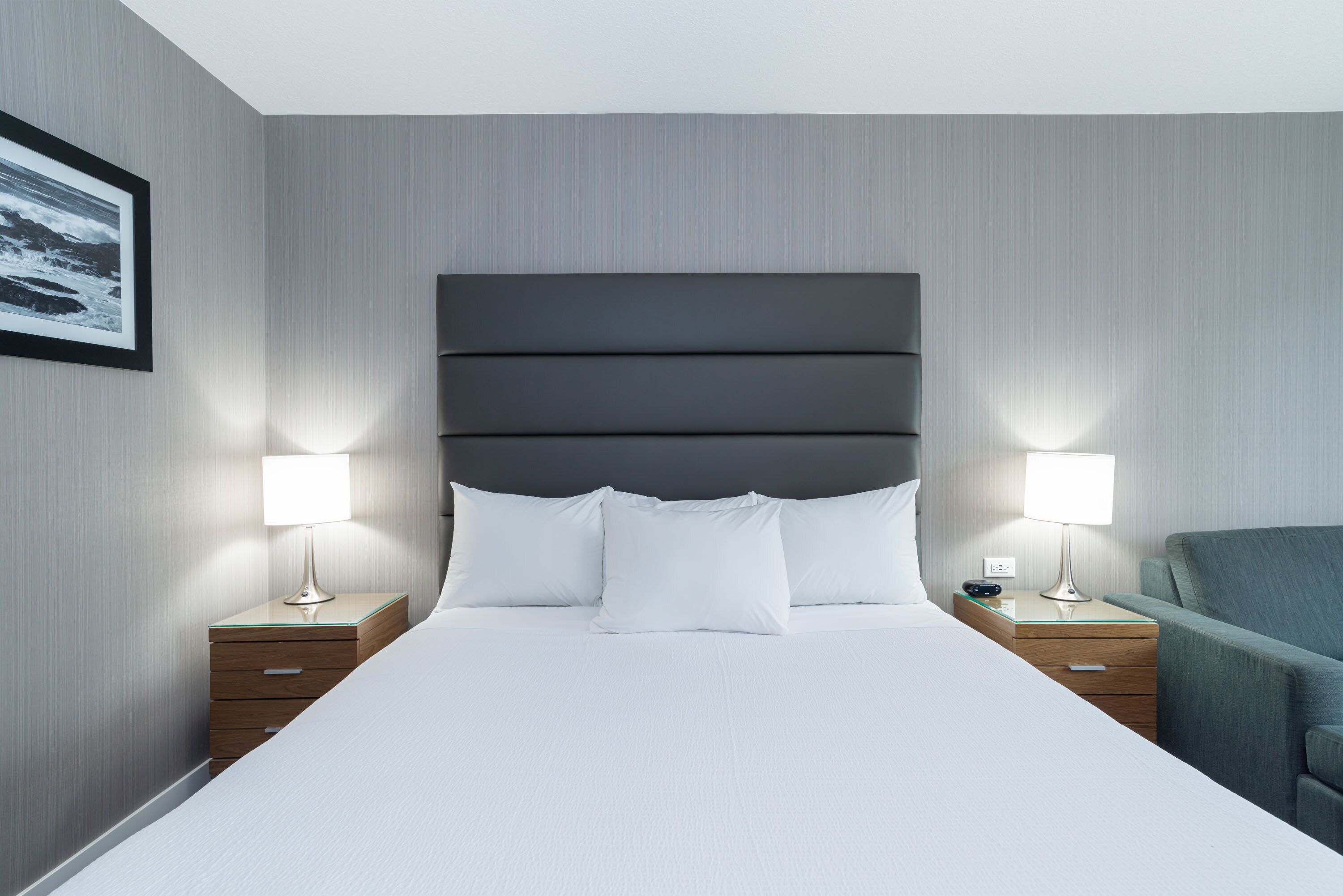 Une chambre d’hôtel moderne avec un grand lit, deux tables de chevet avec des lampes et un fauteuil à revêtement gris.