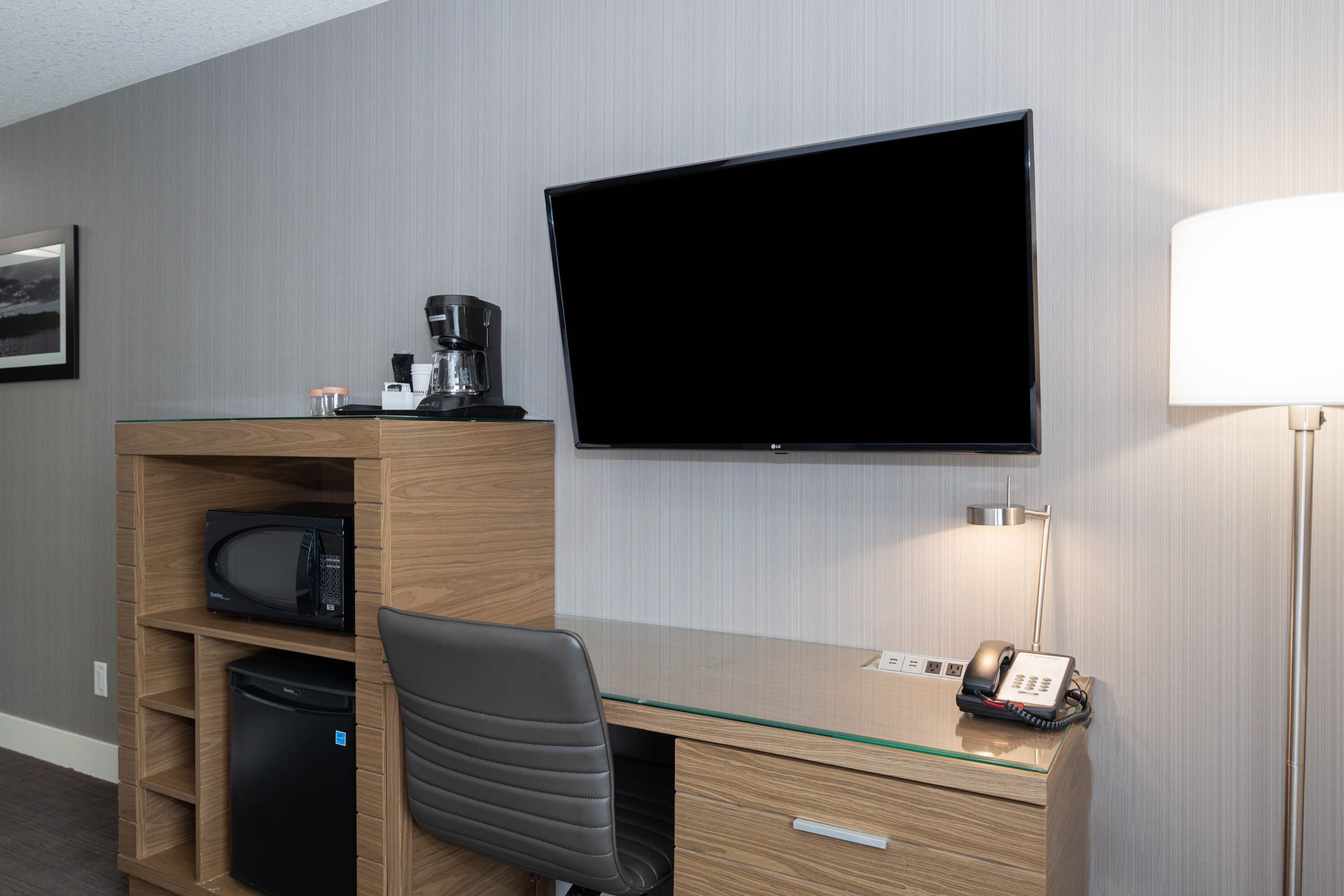 A hotel room with a wooden desk, a flat-screen TV, a microwave, a coffee maker, a telephone, and a chair.