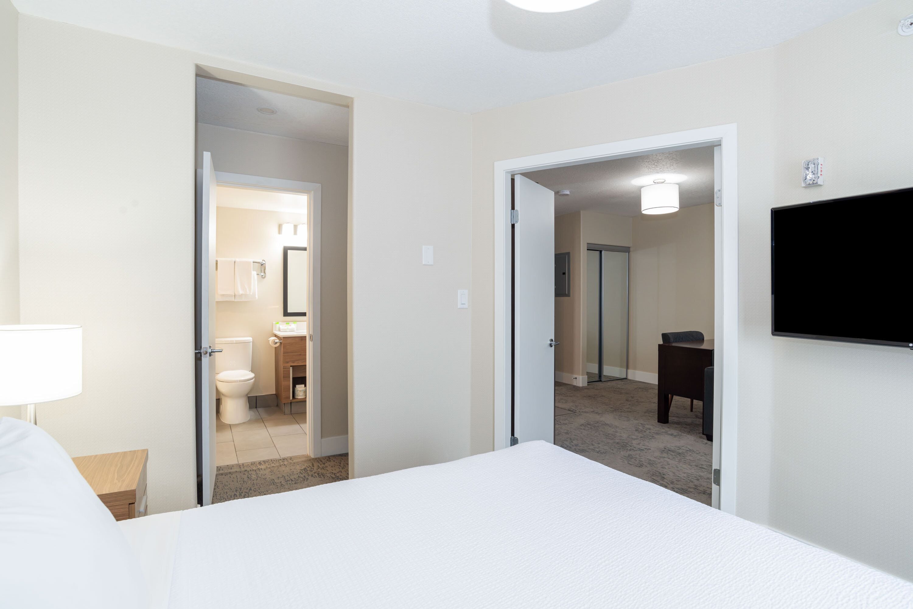 Habitación de hotel con cama, televisión montada en la pared, baño con inodoro y lavamanos, y piso alfombrado.