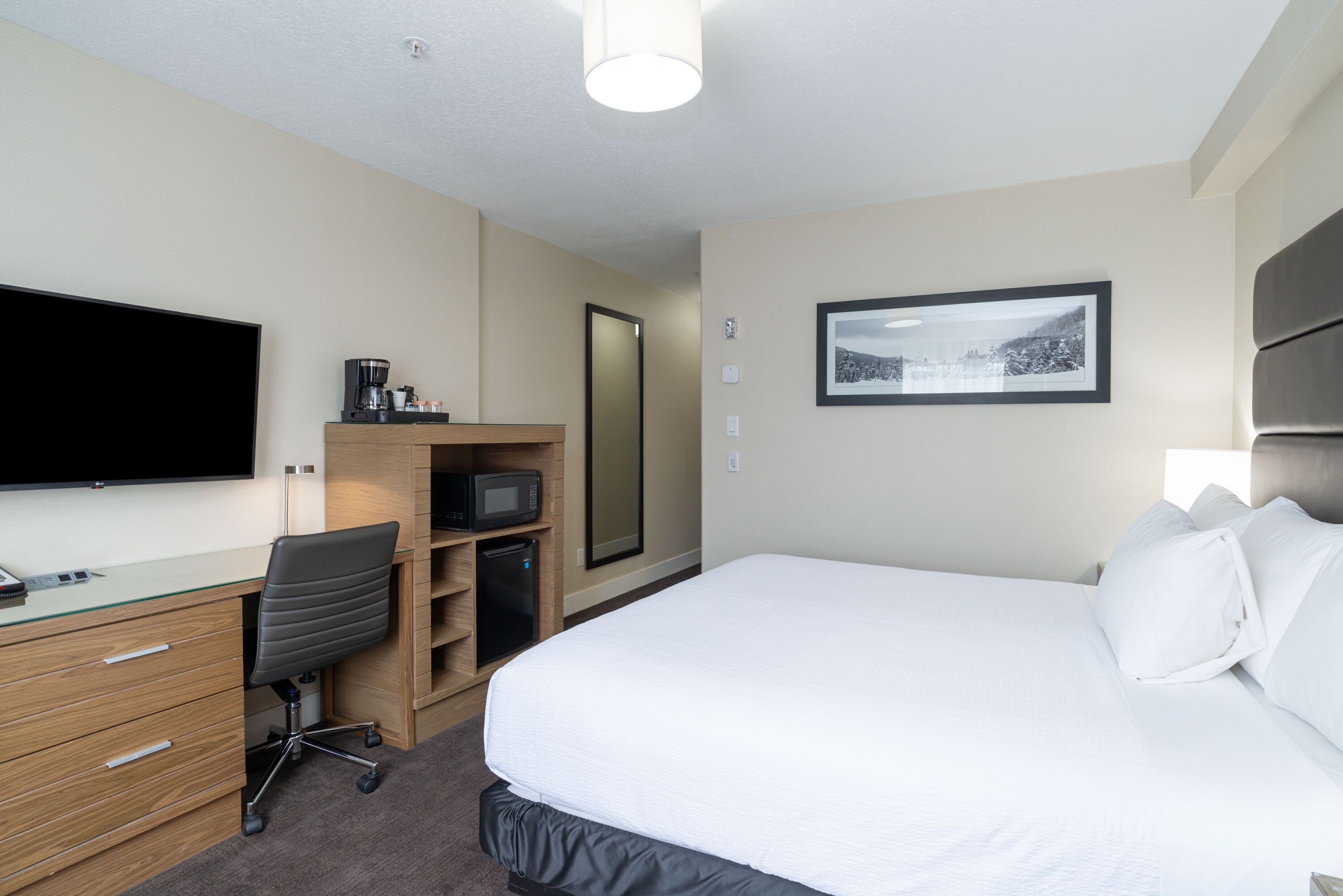 A hotel room with a bed, a desk with a computer, a chair, a TV, a microwave, and a framed picture on the wall.