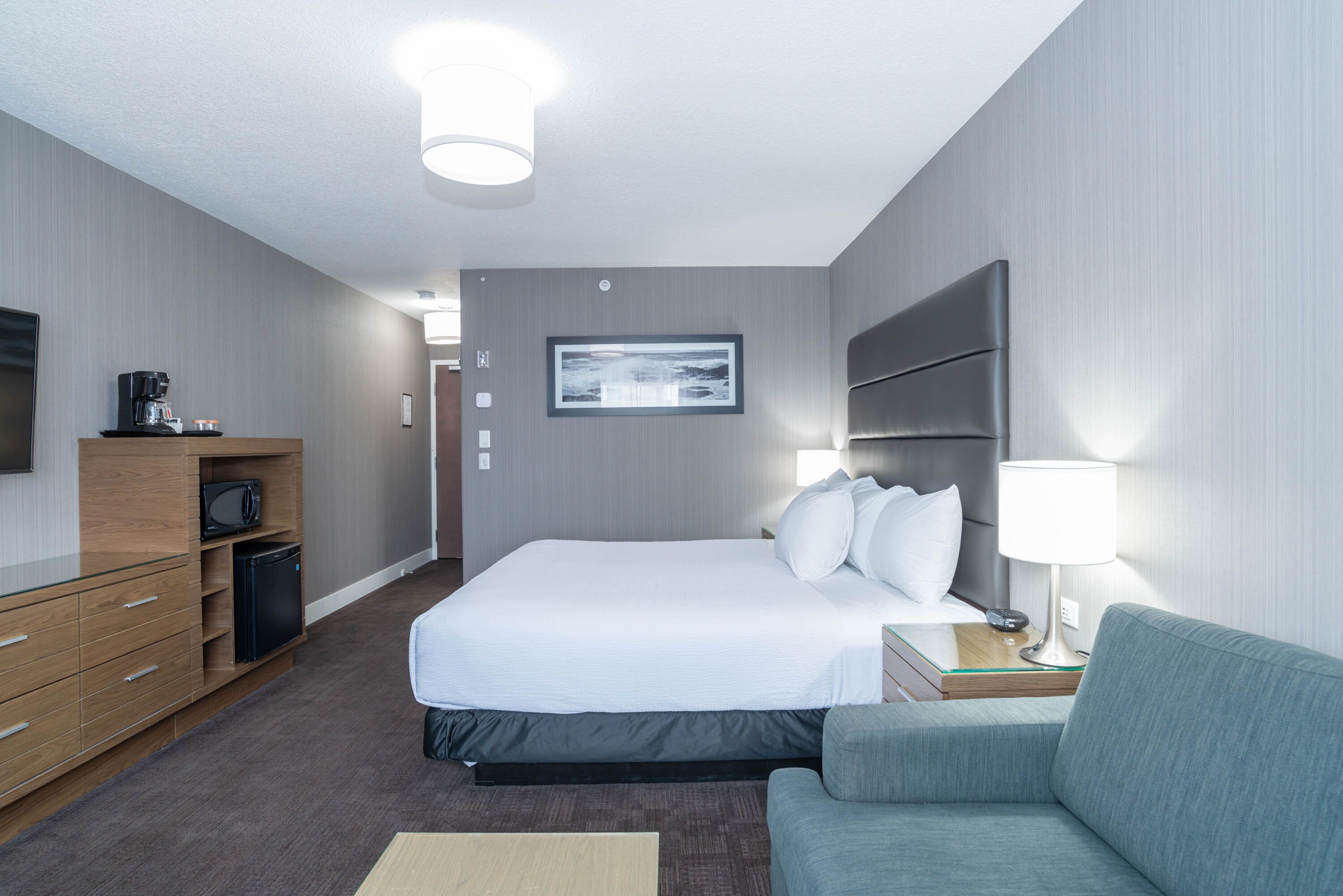 A hotel room with a bed, a desk with a lamp, a sofa, a microwave, a coffee maker, and a framed picture on the wall.