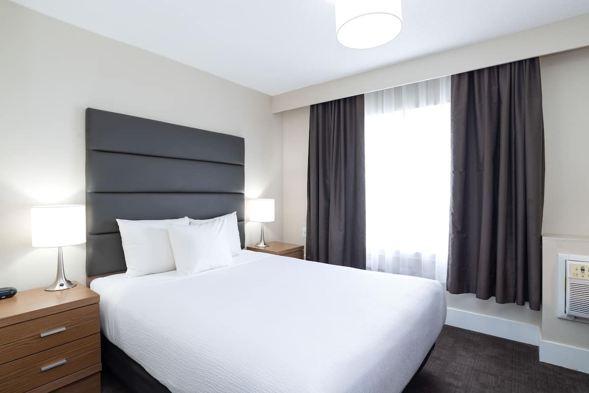 A hotel room with a large bed, two bedside tables with lamps, a modern headboard, and a large window with curtains.