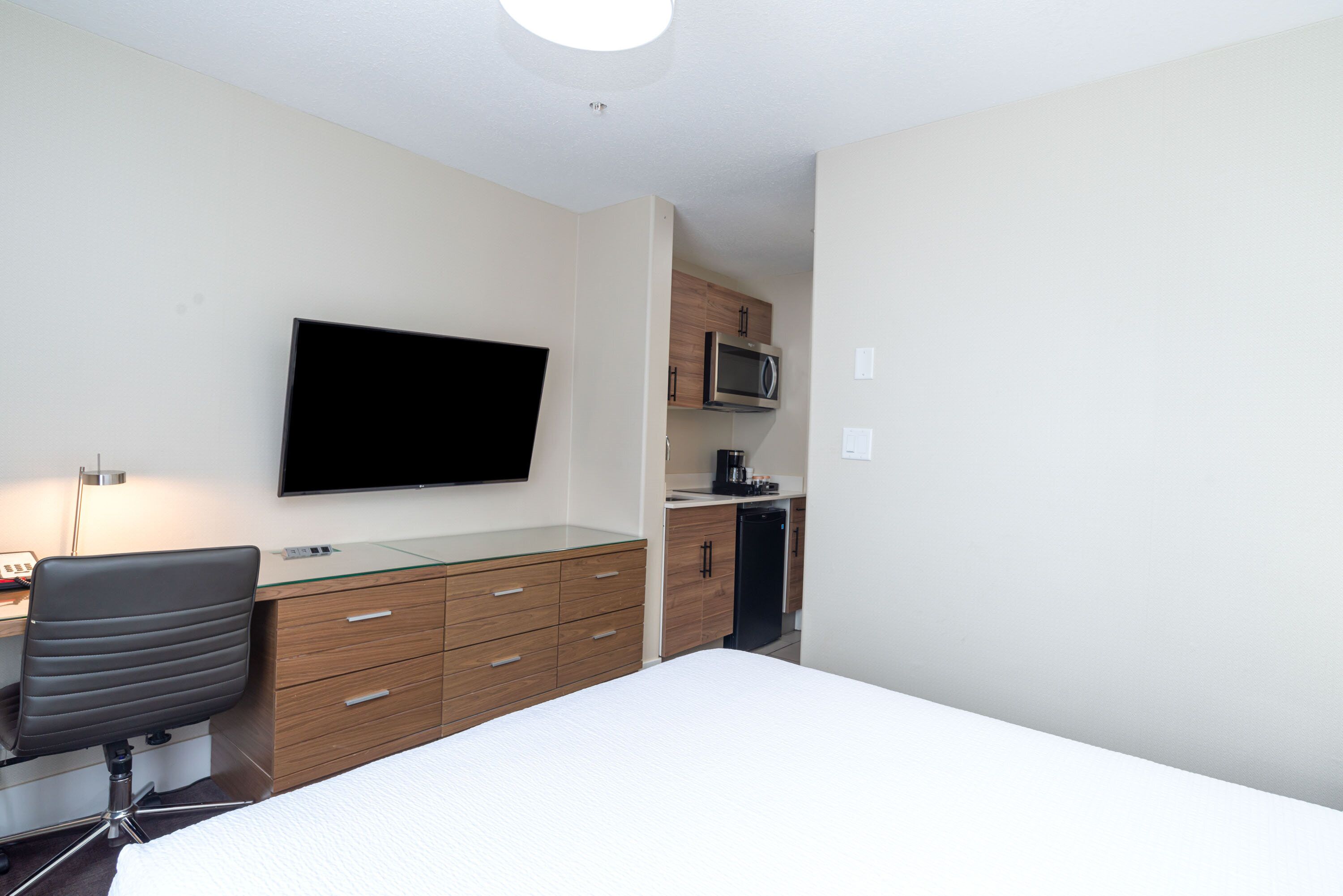 Una habitación pequeña con cama, escritorio con silla, televisor de pantalla plana, microondas y cafetera.
