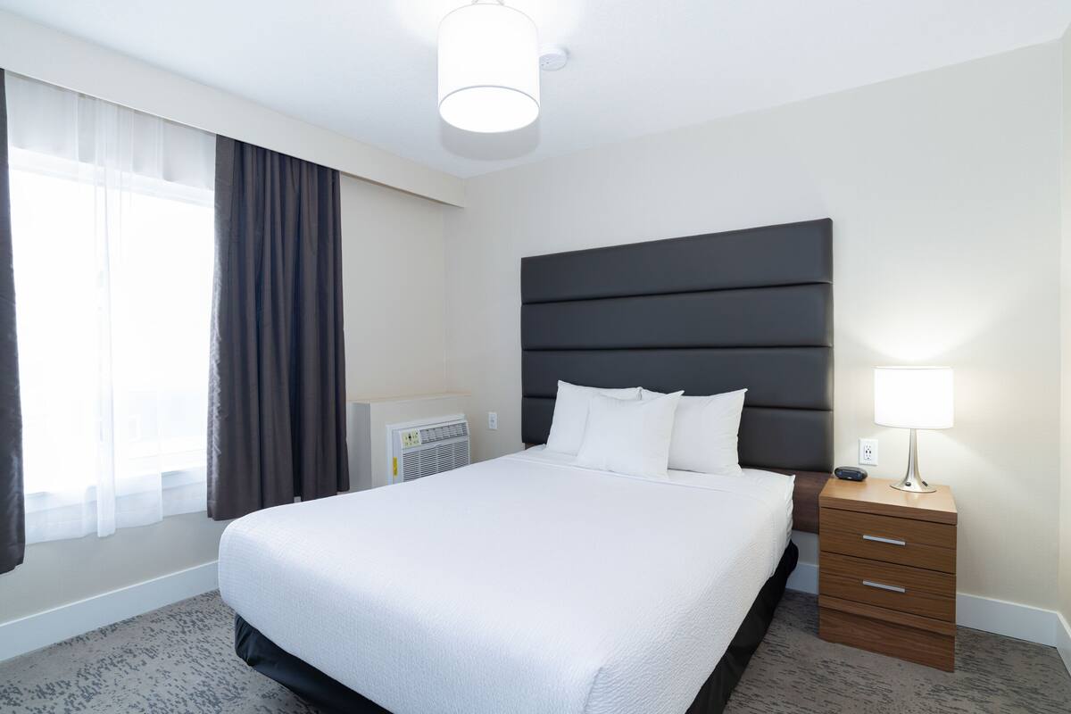 A neatly made bed with a dark headboard, a bedside lamp, and a small wooden nightstand with drawers.