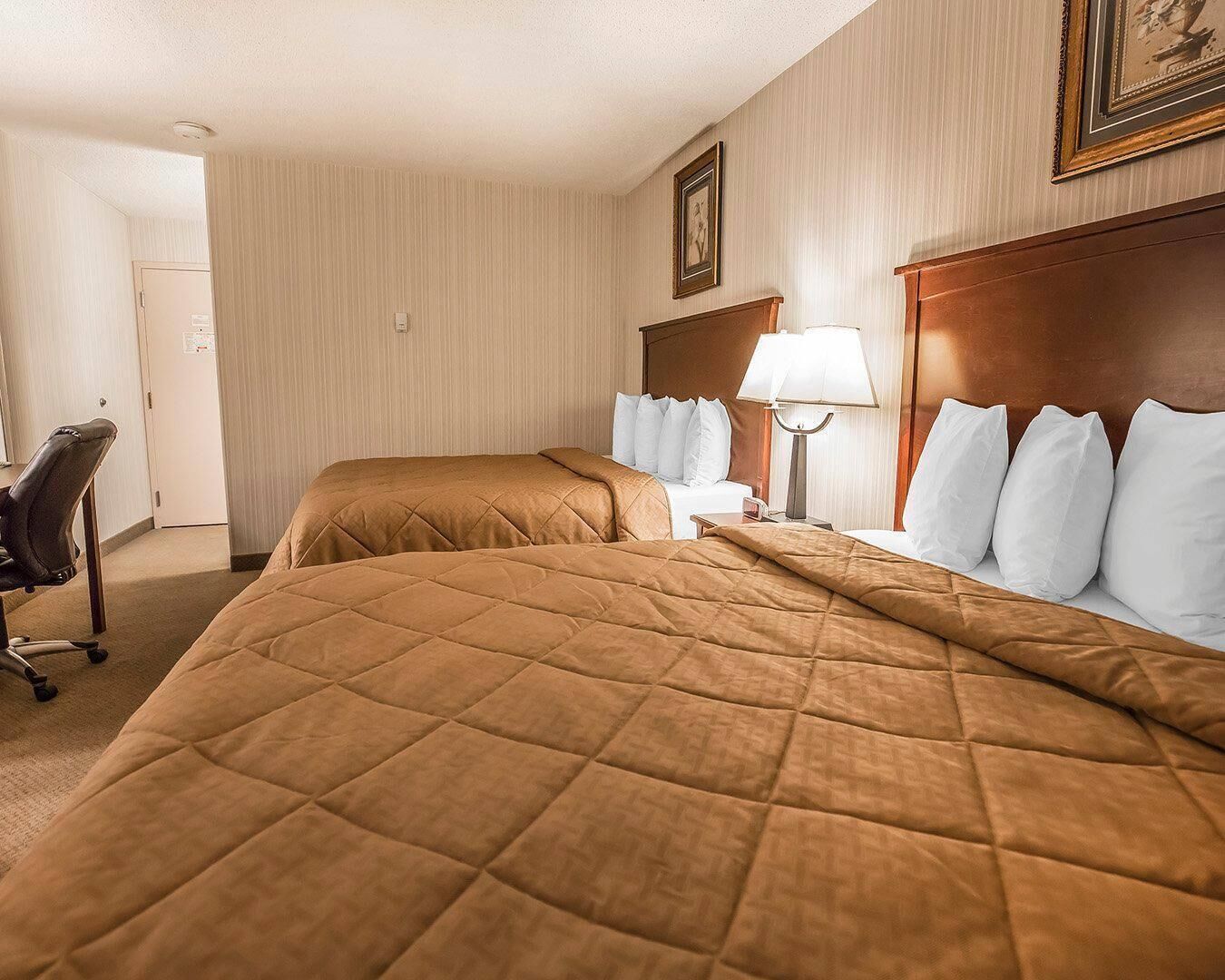 A hotel room with two beds, a desk, a chair, and a lamp.