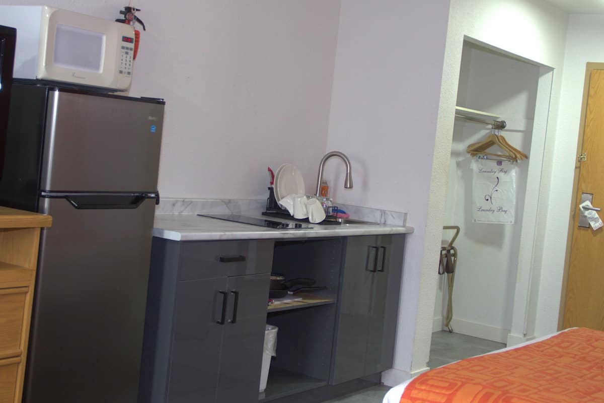 Une cuisine compacte équipée d’un réfrigérateur, d’un four à micro-ondes, d’un évier et d’un petit plan de travail. On y trouve une porte en bois avec une pancarte, une commode en bois et un lit recouvert d’une couverture orange.