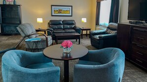 A hotel room with a seating area featuring two blue armchairs, a small round table with a vase of pink flowers, a sofa, and a television on a cabinet.