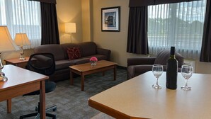A hotel room with a sofa, a small table, a desk, and a chair. There are two windows with curtains, a lamp, and a framed picture on the wall.