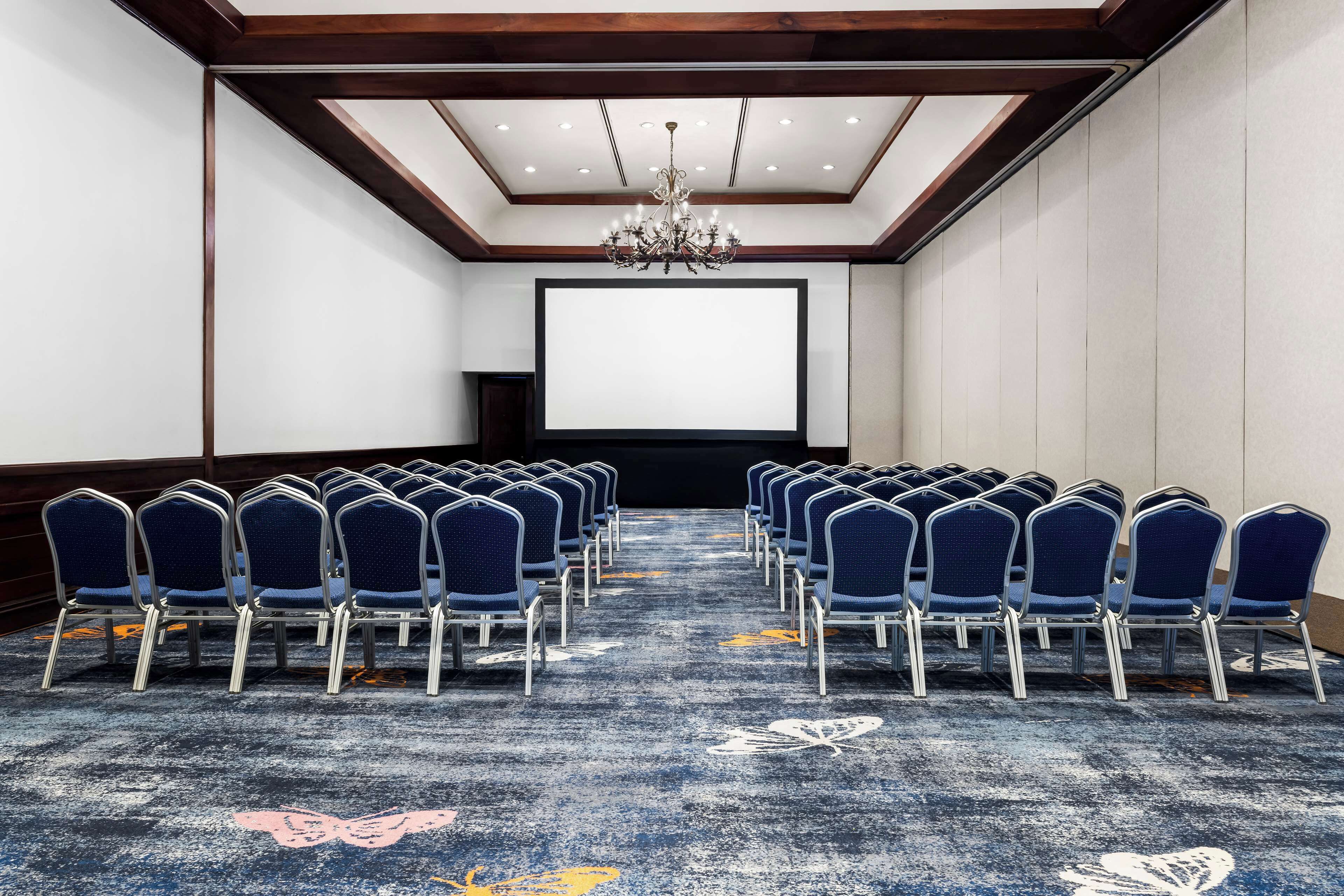 Een congreszaal met rijen blauwe stoelen, een groot scherm en een kroonluchter.