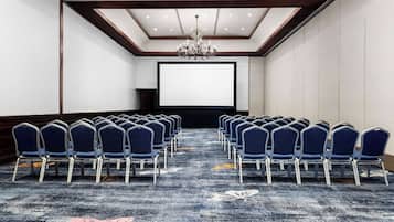Un salón de conferencias con filas de sillas azules, una pantalla grande y una araña.