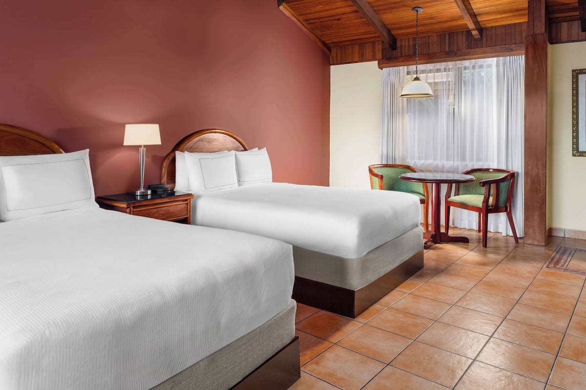 A hotel room with two beds, a small table with chairs, a lamp, and a window with curtains.