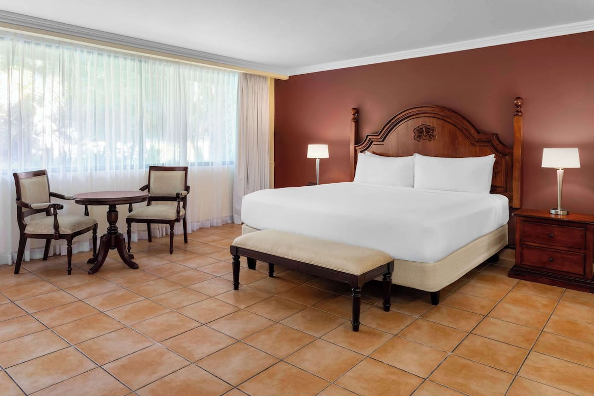 A hotel room with a large bed, a small table with chairs, and a bench.