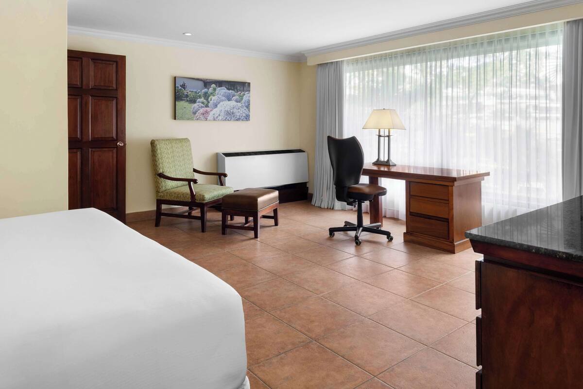A hotel room with a bed, a desk, a chair, a lamp, and a window with curtains.
