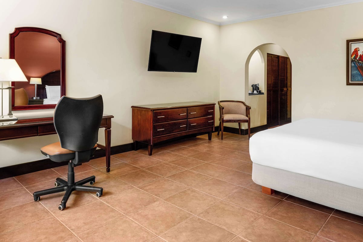 A hotel room with a bed, a desk with a chair, a dresser, a TV mounted on the wall, and a painting on the wall.