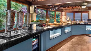 A buffet area with various food items, a refrigerated display, and a view of tropical plants through large windows. - DoubleTree by Hilton Cariari - San Jose Costa Rica (La Asunción)