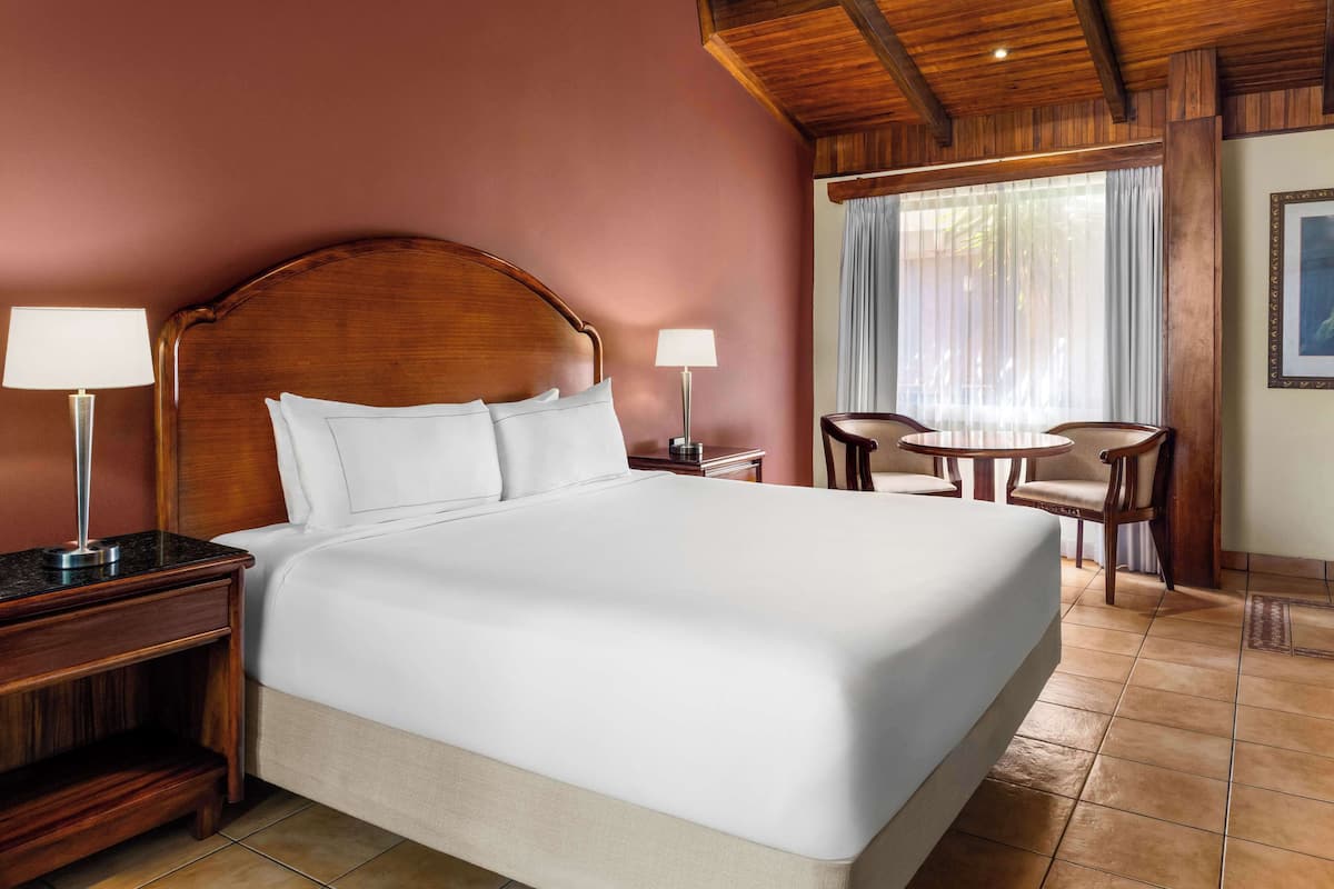 A hotel room with a large bed, two bedside tables with lamps, a small round table with chairs, and a large window with curtains.