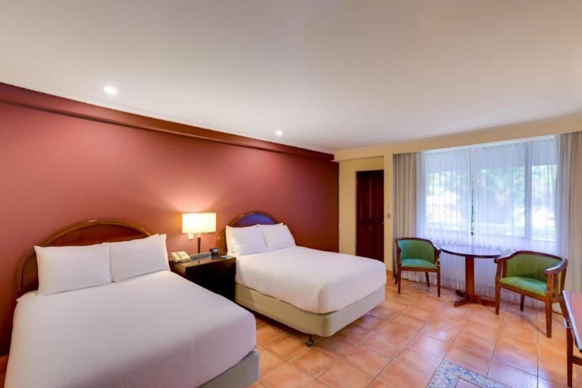 A hotel room with two beds, a desk, chairs, and a window with curtains.