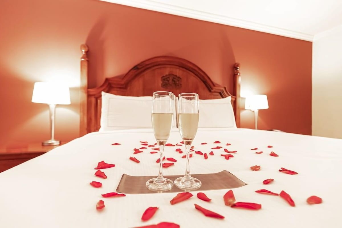 A hotel room with a bed, two bedside lamps, and two champagne glasses on the bed.