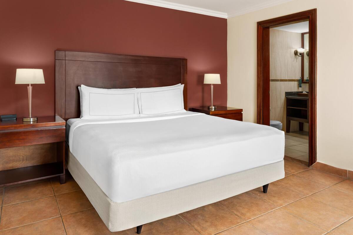 A hotel room with a large bed, two bedside tables with lamps, a wooden headboard, and a bathroom visible through an open door.