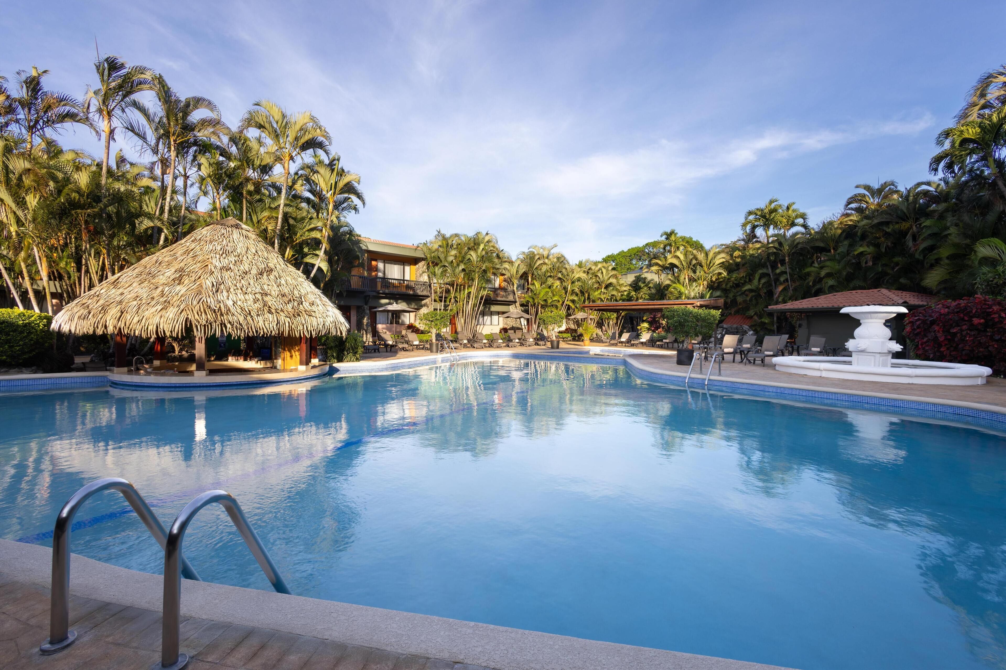 A swimming pool with a thatched-roof structure, palm trees, and a white fountain.