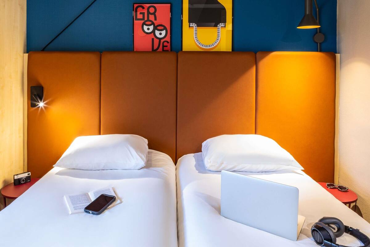 Une chambre d’hôtel avec deux lits, un ordinateur portable sur l’un des lits, un casque audio sur l’autre, et une petite table avec une lampe et une paire de lunettes de soleil.