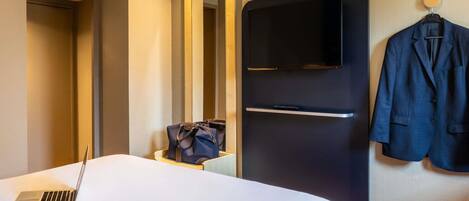 A hotel room with a bed, a desk with a laptop and headphones, a TV, a coat hanging, and a mirror.