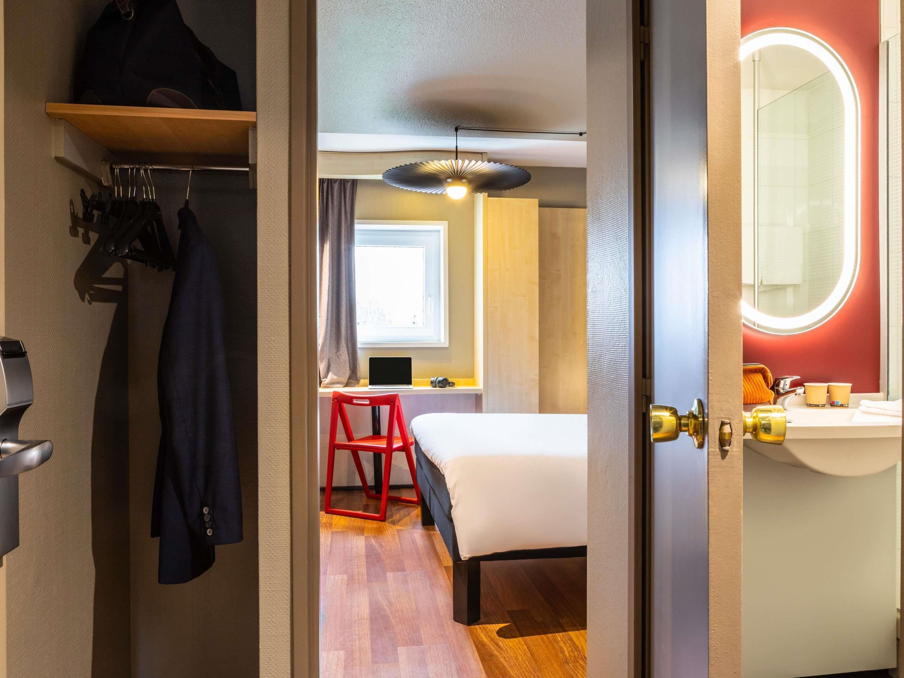Une chambre d’hôtel avec un lit, un bureau avec un ordinateur portable, une chaise rouge et une salle de bain avec un miroir et un lavabo.