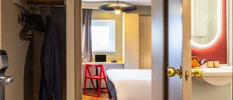 Habitación de hotel con cama, escritorio con una computadora portátil, una silla roja y un baño con espejo y lavamanos.