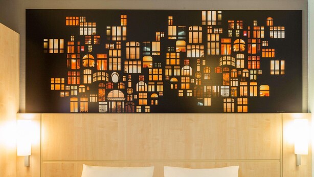 Hotelkamer met grote wanddecoratie van verlichte ramen, een houten hoofdeinde en twee bedlampen.
