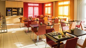 Une salle à manger d’hôtel avec de nombreuses tables et chaises, un coin cuisine équipé d’appareils électroménagers et de grandes fenêtres avec des rideaux.