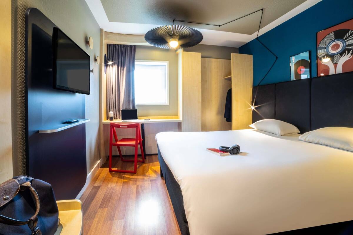 Une chambre d’hôtel avec un lit, un bureau avec un ordinateur portable, une chaise rouge, un téléviseur fixé au mur et un luminaire au plafond.