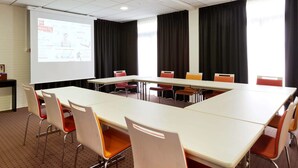 Une salle de conférence équipée d’une longue table, de chaises, d’un écran de projection et d’un présentateur.