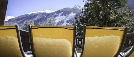 Three yellow chairs with metal frames, positioned outdoors with a view of snow-capped mountains and trees in the background.