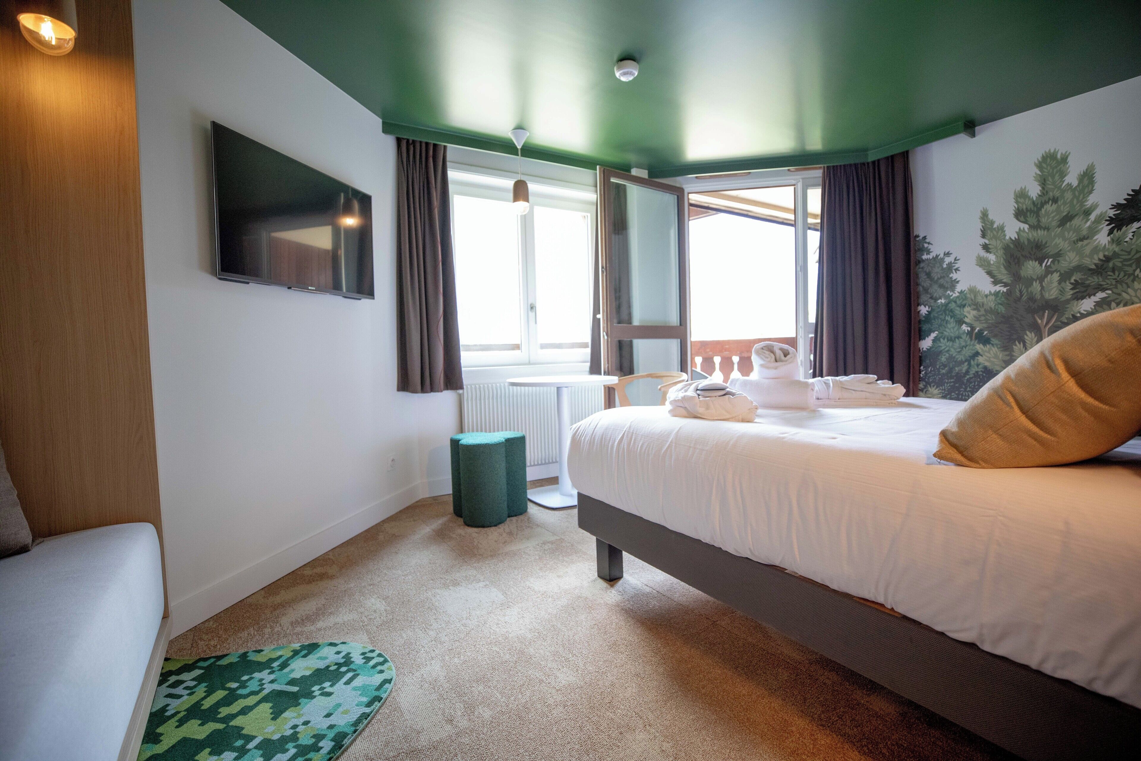 A bedroom with a bed, a TV mounted on the wall, a window with curtains, and a radiator.