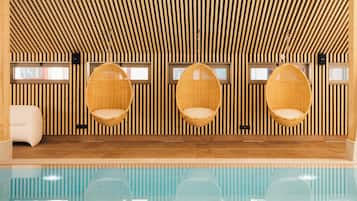 Un área de piscina interior moderna con paredes de listones de madera, tres sillas de mimbre y un sofá blanco.