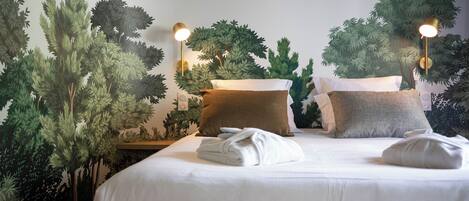 A neatly made bed with white linens and a brown pillow, a wall-mounted lamp, and a large artificial tree mural.