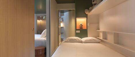 A bedroom with a bed, a bedside table, a nightstand, a wardrobe, and a teddy bear on a shelf.