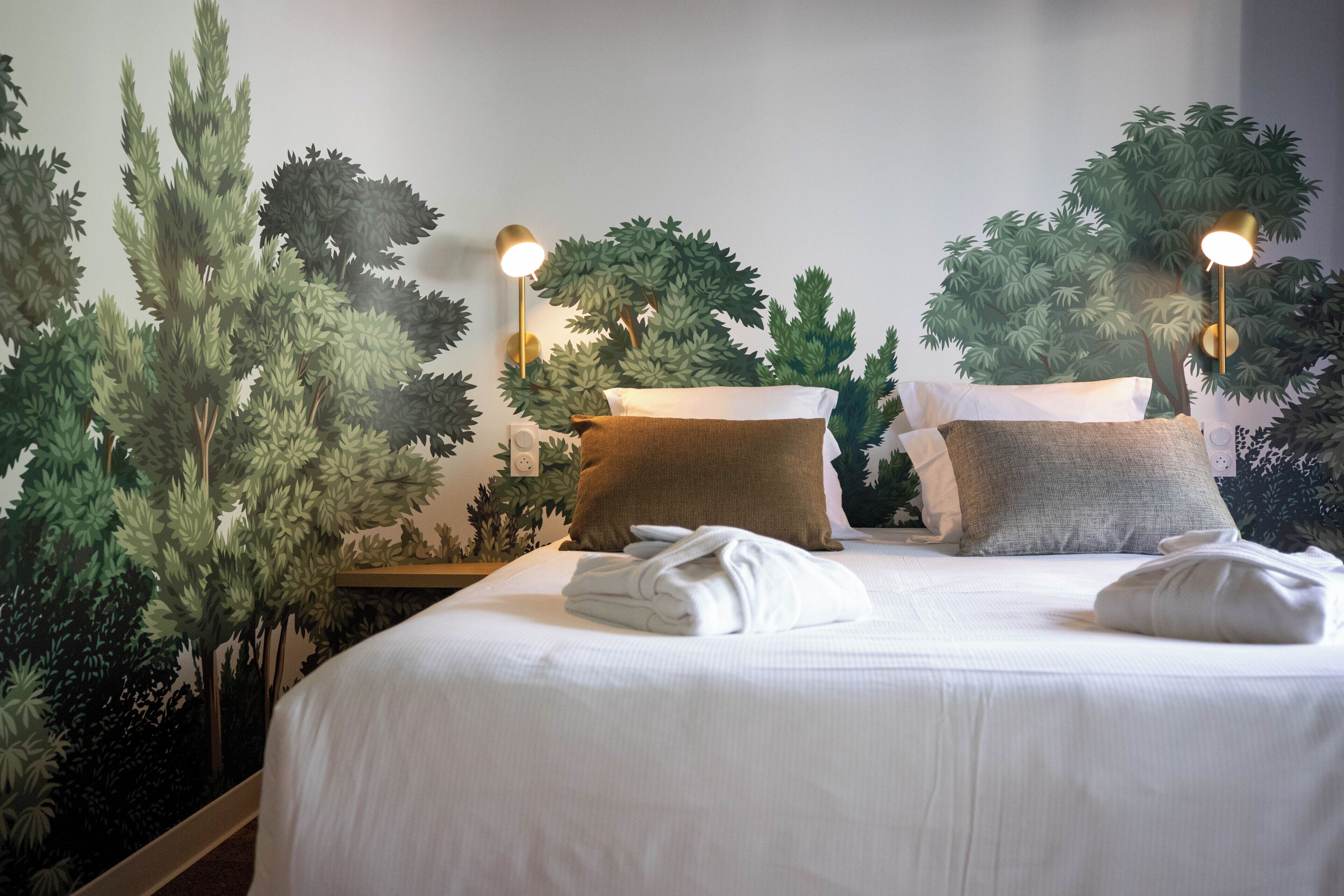 A neatly made bed with white linens and a brown pillow, a wall-mounted lamp, and a large artificial tree mural.