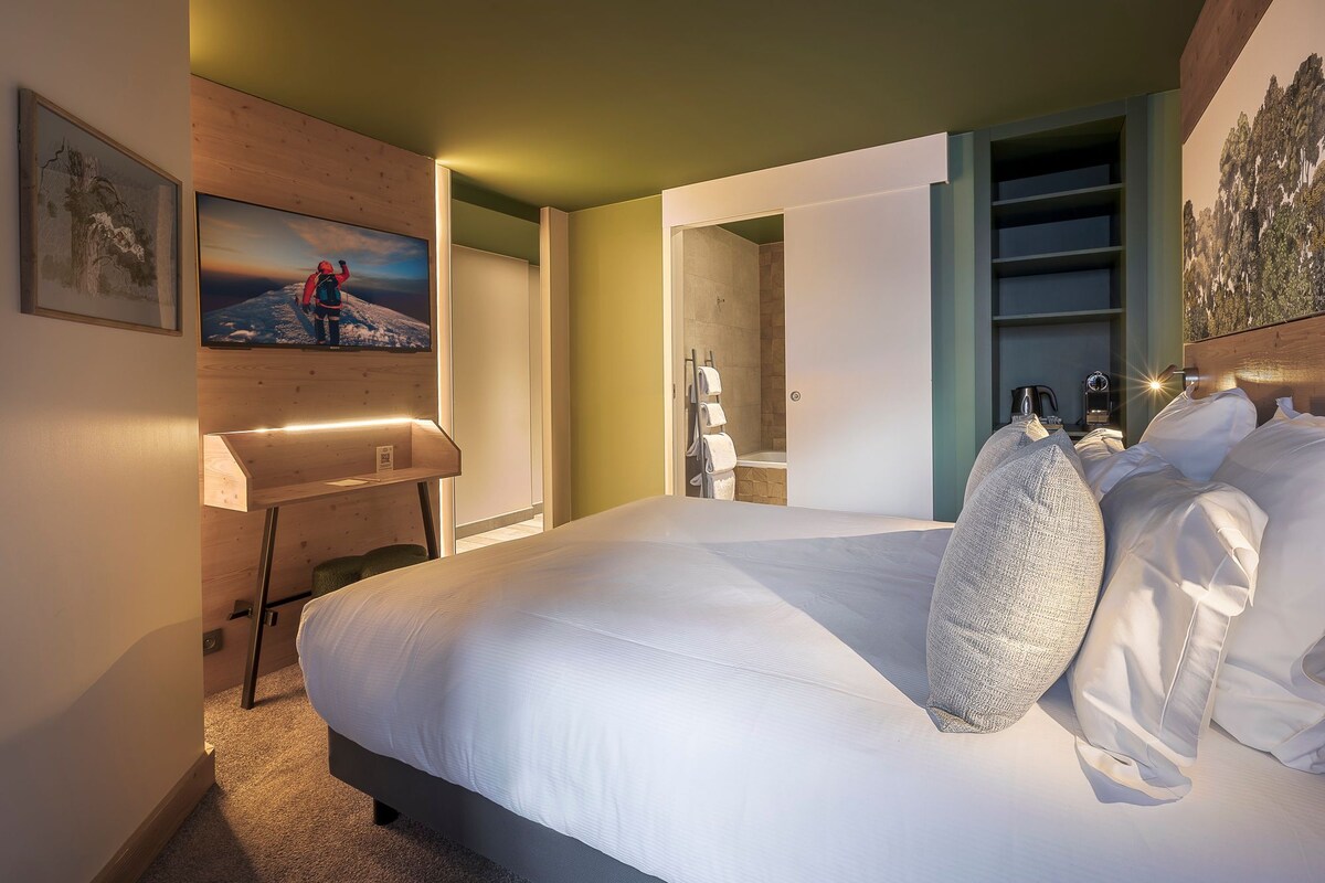 Una habitación de hotel moderna con una cama grande, un escritorio de madera, zona de ducha y un cuadro en la pared.