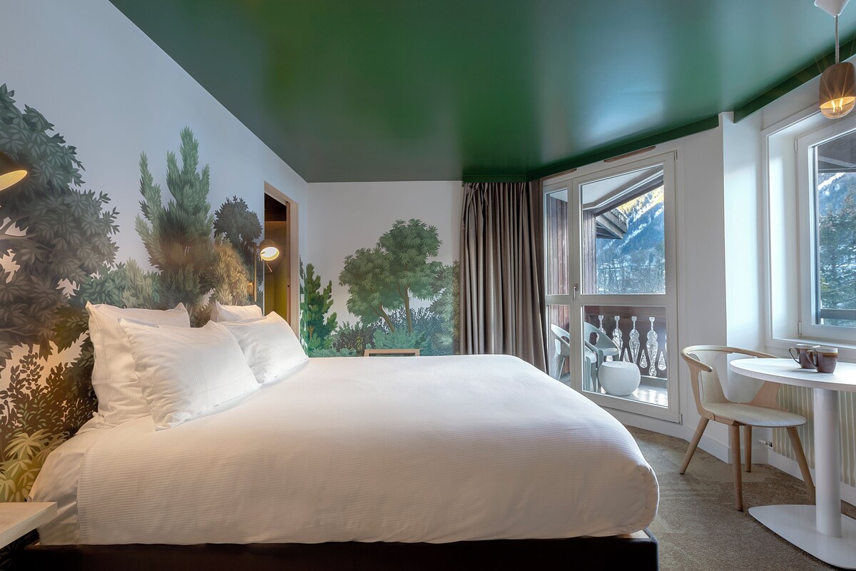 Habitación de hotel con una cama grande, zona de comedor con una mesa y sillas, y vista a árboles por la ventana.