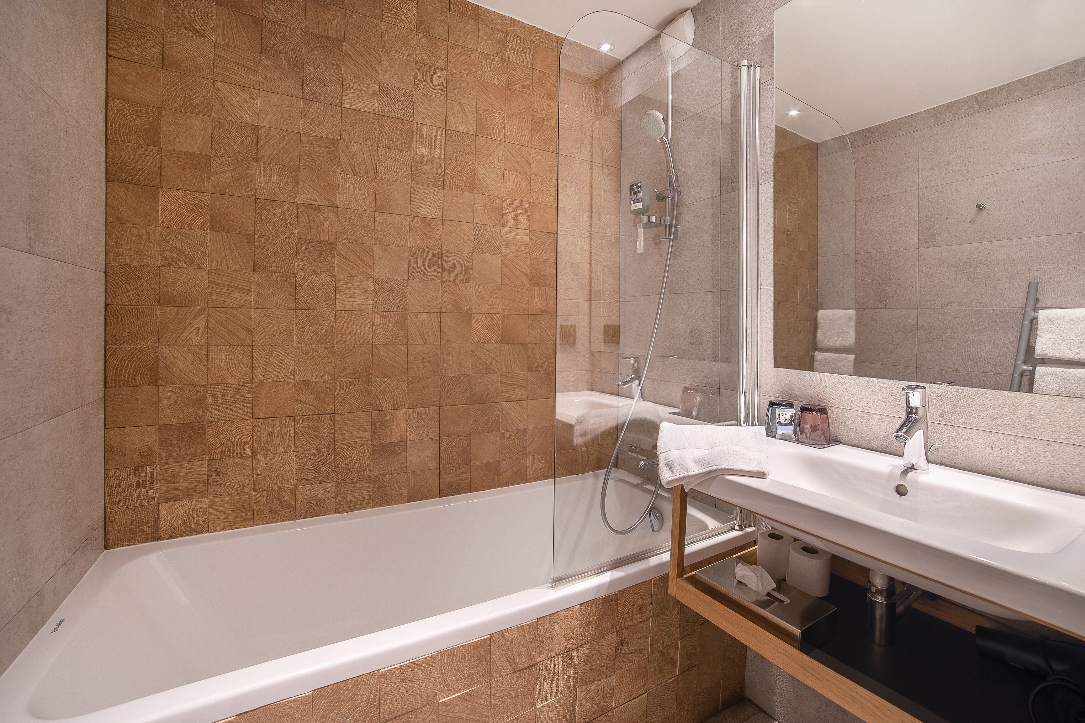 A modern bathroom with a wooden wall, a glass-enclosed shower, a white bathtub, and a sink with a mirror.