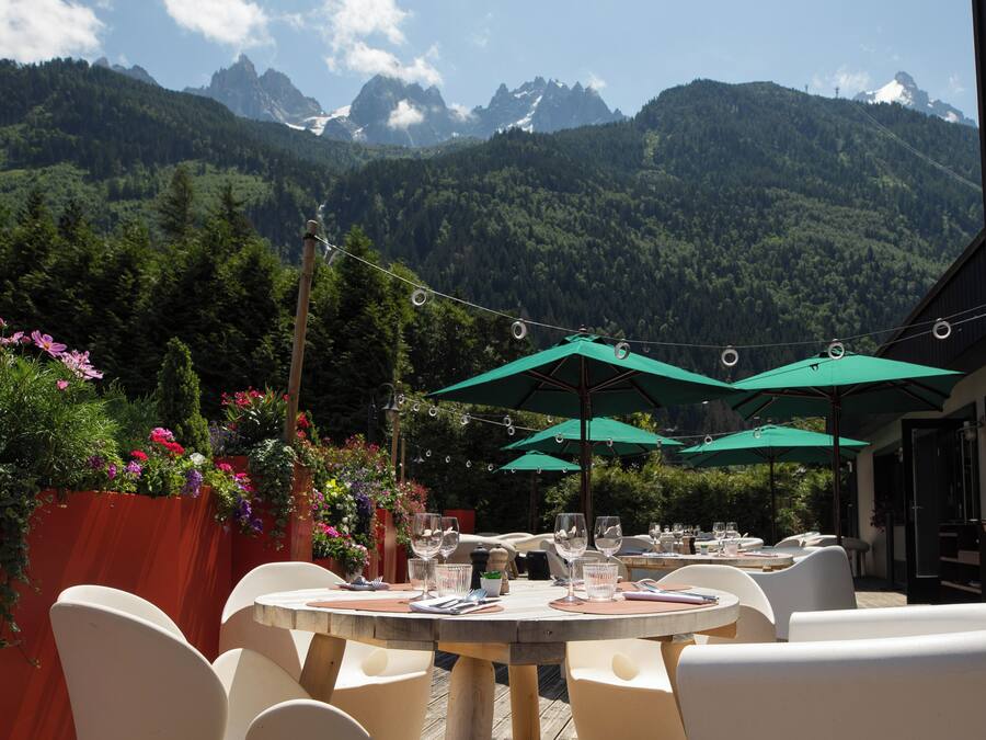 An outdoor dining area with round tables and white chairs, surrounded by greenery and a mountain backdrop.