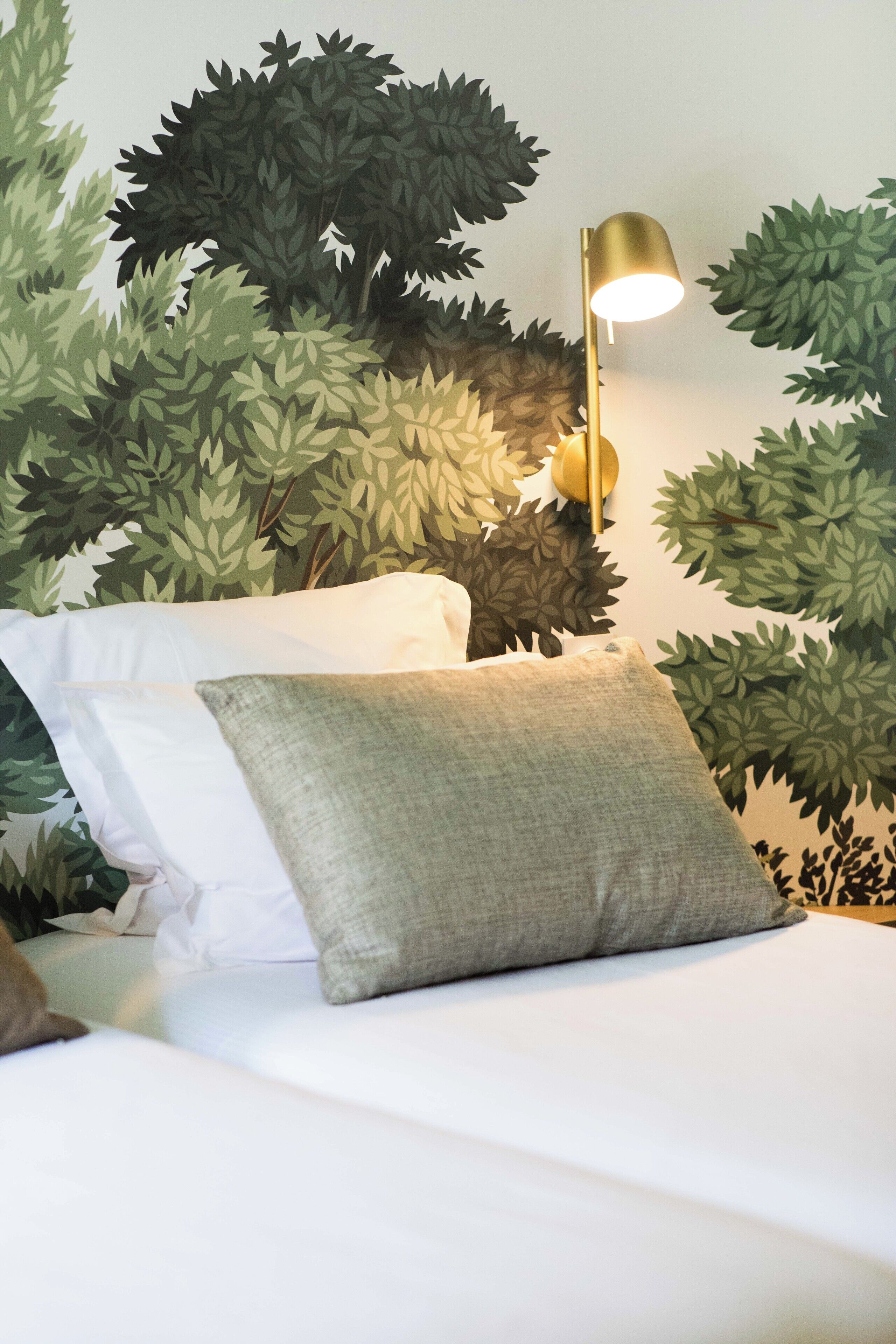 A bed with white linens and a gray pillow, a gold lamp, and a wall with a leafy mural.