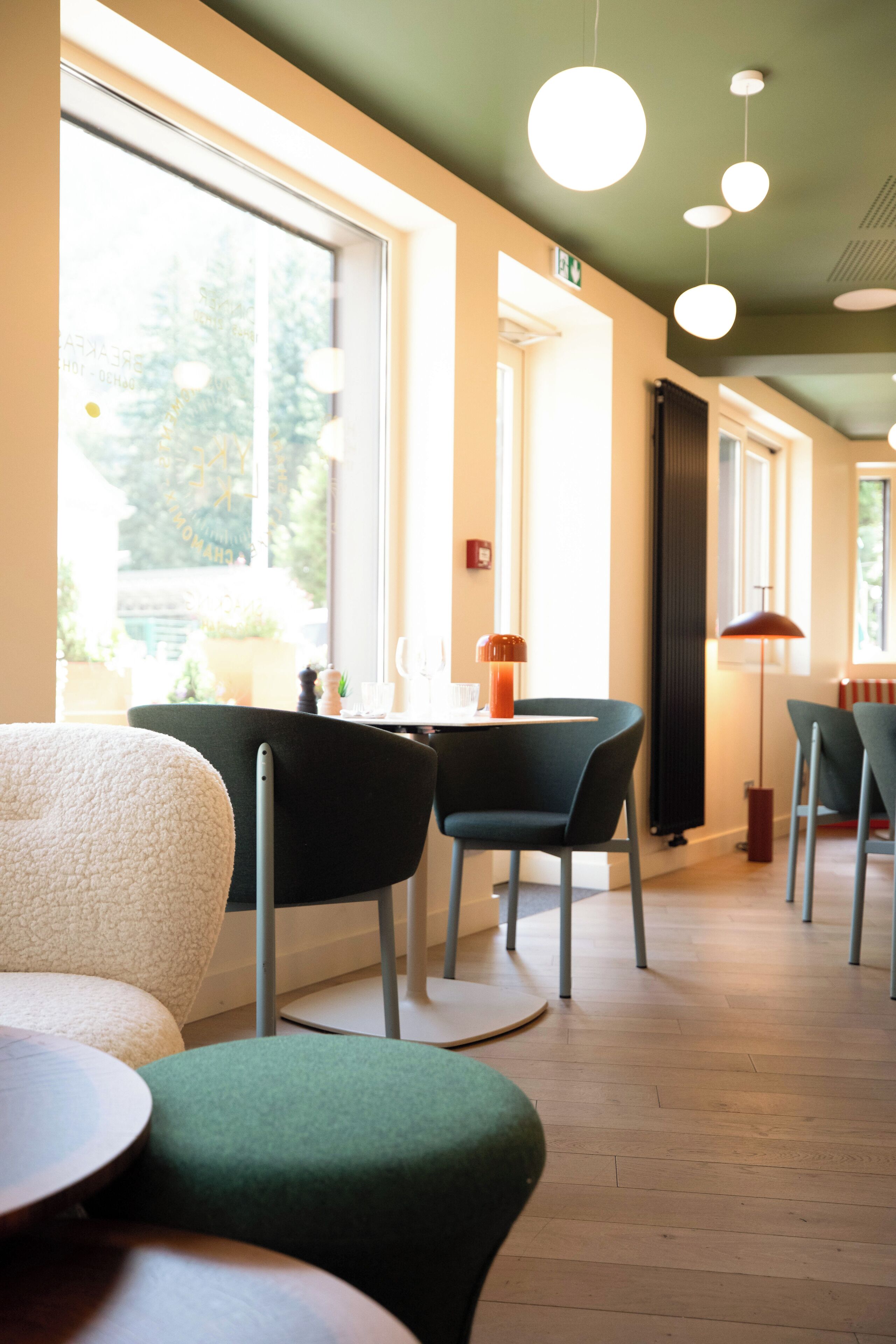 A modern interior with a dining area featuring round tables and chairs, large windows providing natural light, and a visible exit sign.